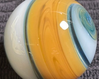 May include: A glass marble featuring a swirling design in white, gold, and blue. The spherical marble has a polished surface. The colours merge in a spiral pattern, producing a distinctive and appealing look.