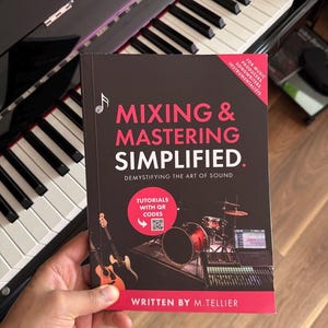 May include: A book titled "MIXING & MASTERING SIMPLIFIED" with the subtitle "DEMYSTIFYING THE ART OF SOUND." The cover features musical notes, a drum set, a guitar, and a mixing board. The book is held near a piano keyboard.