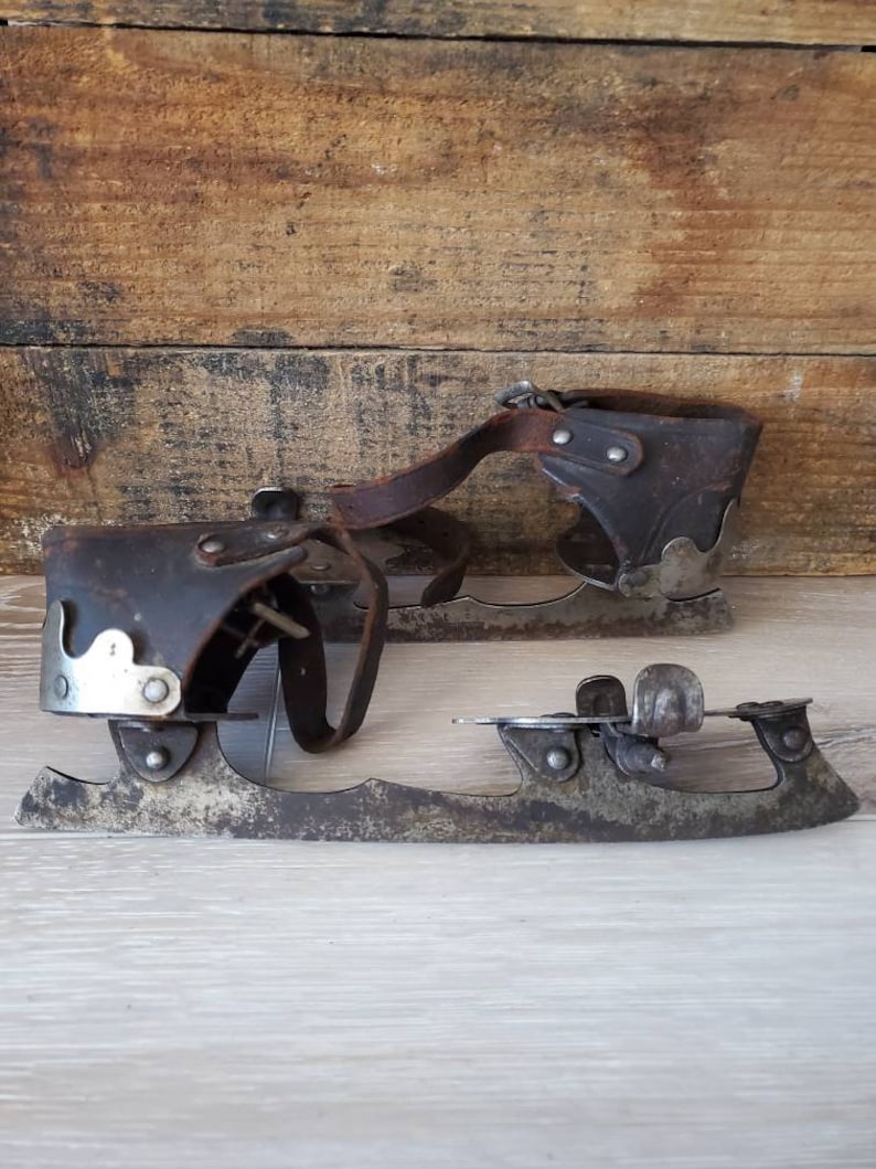 May include: A pair of vintage ice skates, featuring leather straps and metal blades. The skates show signs of wear and age, with a rustic aesthetic. The blades are attached to the leather boots with rivets, showcasing a classic design.
