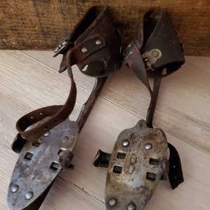 May include: A pair of vintage ice skates, featuring metal blades and leather straps. The skates have a worn, aged look. The blades are secured to the leather boot holders with rivets. These skates are designed to be strapped onto boots for ice skating.