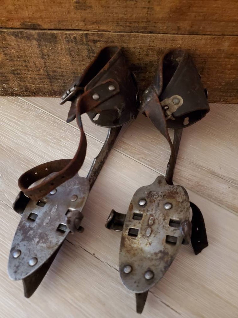 May include: A pair of vintage ice skates, featuring metal blades and leather straps. The skates have a worn, aged look. The blades are secured to the leather boot holders with rivets. These skates are designed to be strapped onto boots for ice skating.
