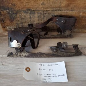 May include: Vintage ice skates with leather heel and clamp toe design. The skates are size 8 1/2 with a 25.4 cm blade. The skates are displayed on a wooden surface. The tag reads "JBC COLLECTION".