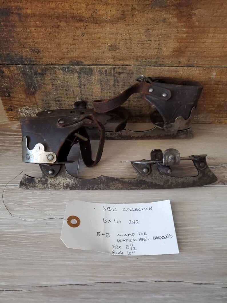 May include: Vintage ice skates with leather heel and clamp toe design. The skates are size 8 1/2 with a 25.4 cm blade. The skates are displayed on a wooden surface. The tag reads "JBC COLLECTION".
