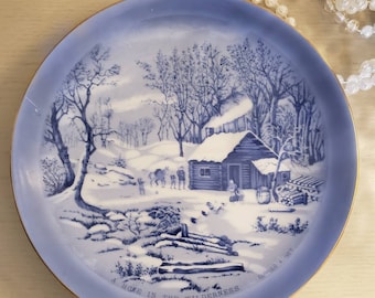 May include: A blue and white ceramic plate with a gold rim depicts a winter scene of a cabin in the woods. The plate reads "A Home in the Wilderness" and is signed "Carpenter &  Wes."