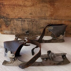 May include: A pair of vintage ice skates featuring leather straps and brown leather ankle supports. The metal blades show signs of wear. The skates are displayed on a light wood surface.