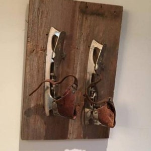 May include: A pair of vintage ice skates mounted on a wooden board. The skates feature silver blades, brown leather straps, and brown leather boot covers. The wood has a weathered, rustic appearance.
