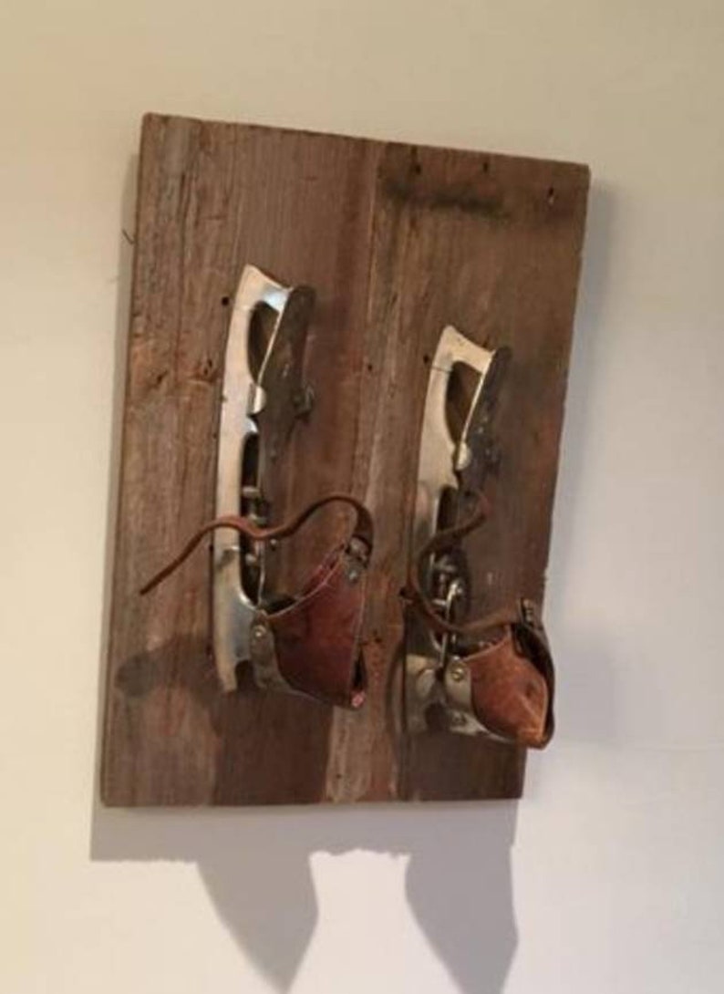 May include: A pair of vintage ice skates mounted on a wooden board. The skates feature silver blades, brown leather straps, and brown leather boot covers. The wood has a weathered, rustic appearance.