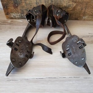 May include: A pair of vintage ice skates, featuring metal blades and leather straps. The skates have a dark, aged metal appearance, showing wear. The leather straps are brown and worn. The skates are displayed on a light wooden surface, suggesting age and use.