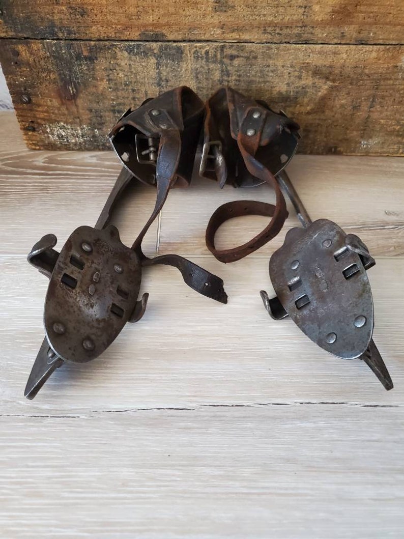 May include: A pair of vintage ice skates, featuring metal blades and leather straps. The skates have a dark, aged metal appearance, showing wear. The leather straps are brown and worn. The skates are displayed on a light wooden surface, suggesting age and use.