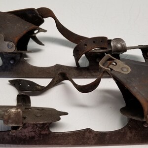 May include: A pair of antique ice skates, with dark brown leather boots and aged metal blades. The skates have leather straps and metal buckles for securing the feet, showcasing a vintage design.