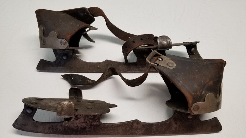 May include: A pair of antique ice skates, with dark brown leather boots and aged metal blades. The skates have leather straps and metal buckles for securing the feet, showcasing a vintage design.