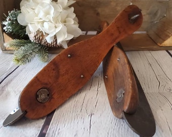 May include: A pair of antique wooden ice skates with metal blades. The skates are brown and have a worn finish. They are sitting on a white wooden surface.