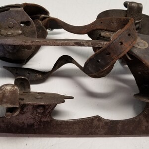 May include: Vintage ice skates, featuring metal blades and leather straps, showing signs of wear and rust. The design includes a curved blade and a mechanism for attaching to boots. A classic, antique item, likely from a bygone era.