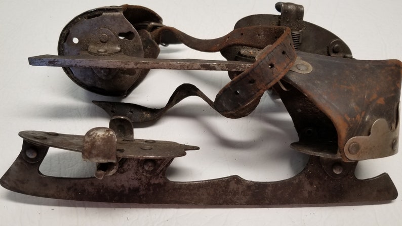 May include: Vintage ice skates, featuring metal blades and leather straps, showing signs of wear and rust. The design includes a curved blade and a mechanism for attaching to boots. A classic, antique item, likely from a bygone era.