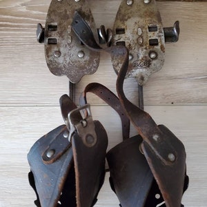 May include: A pair of vintage ice skates. The skates have metal blades with leather straps and boot holders. The metal blades are riveted to the boot holders. The leather straps and boot holders are dark brown. The skates are set against a wooden background.