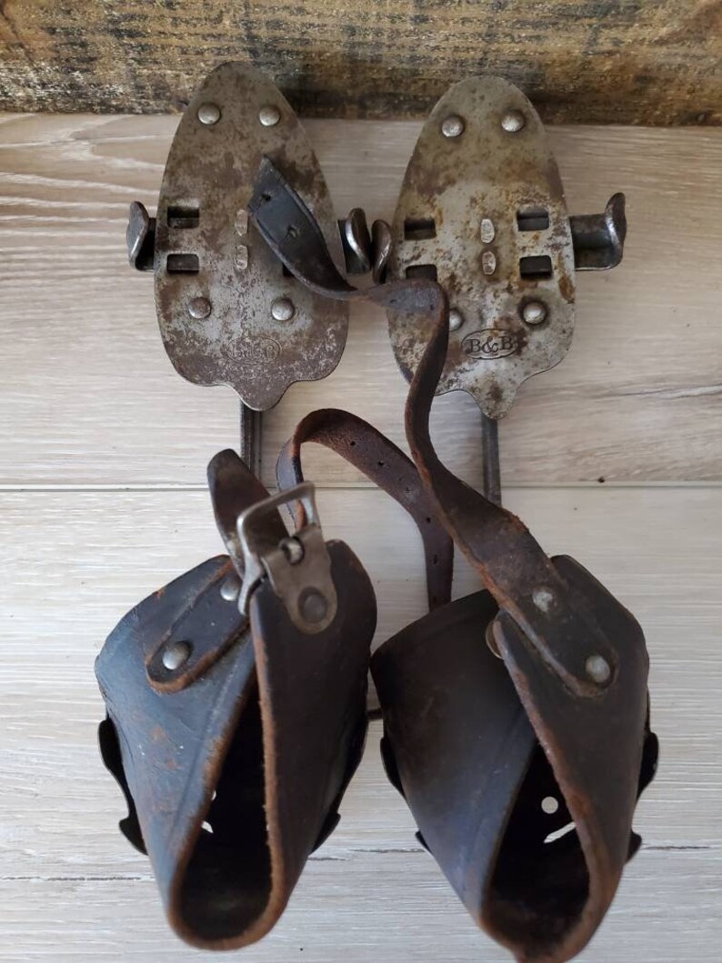 May include: A pair of vintage ice skates. The skates have metal blades with leather straps and boot holders. The metal blades are riveted to the boot holders. The leather straps and boot holders are dark brown. The skates are set against a wooden background.
