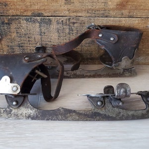 May include: A pair of vintage ice skates, featuring dark brown leather boot holders and metal blades. The skates show signs of age, with wear on the leather and metal parts, giving them a classic look.
