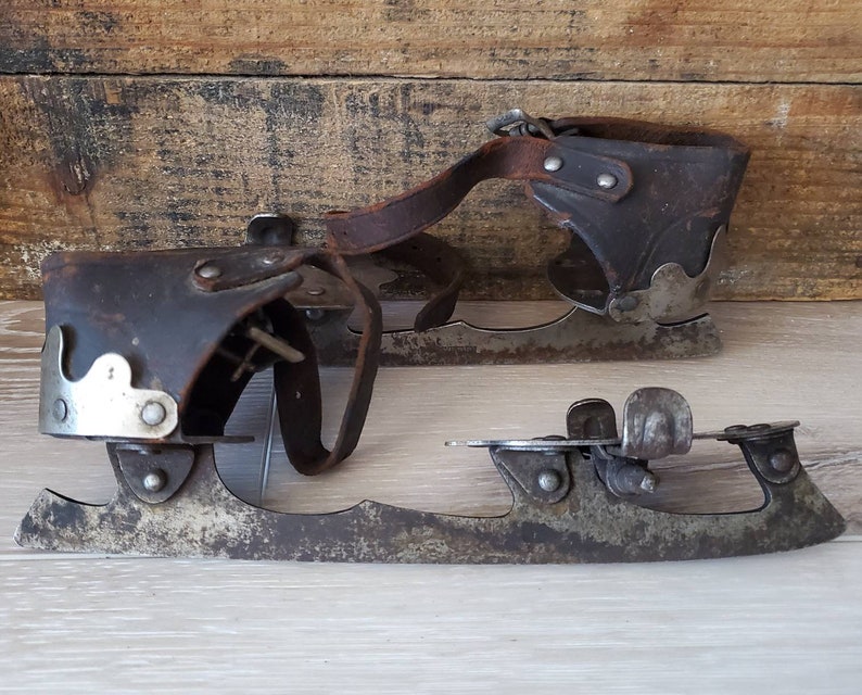 May include: A pair of vintage ice skates, featuring dark brown leather boot holders and metal blades. The skates show signs of age, with wear on the leather and metal parts, giving them a classic look.