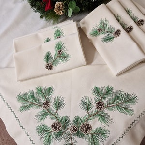 May include: Set of six white linen napkins featuring embroidered green pine branches and brown pine cones. The napkins are displayed on a white tablecloth with a green embroidered border.