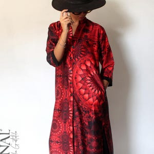 Puede incluir: Un vestido largo abotonado de color rojo y negro con un estampado y un gran diseño circular en la parte delantera. El vestido tiene mangas largas y bolsillos. El texto "SUBILIMINAL by Claudia Gastaldi" está impreso en el lado izquierdo. Se lleva un sombrero negro.