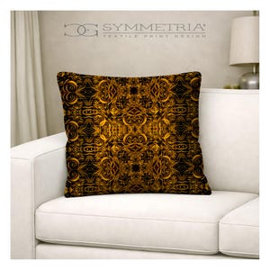 May include: Square decorative pillow with a black and gold ornate design. The pillow features a symmetrical pattern of swirling, floral-like shapes. The pillow is on a white couch. The text "SYMMETRIA TEXTILE PRINT DESIGN" is at the top.