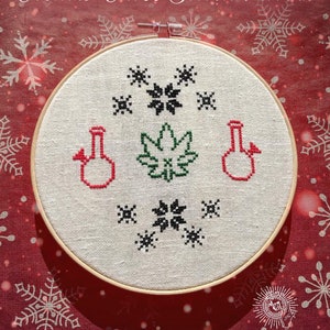 May include: A white embroidery hoop with a cross stitch pattern of a marijuana leaf, two bongs, and snowflakes. The hoop is on a red background with white snowflakes.