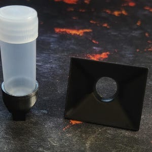 Op de afbeelding: Een doorzichtige plastic buis met een zwarte basis en een zwart vierkant met een rond gat in het midden.