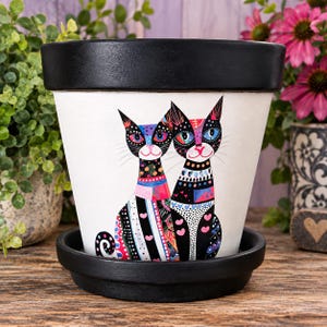 May include: A white ceramic plant pot with a black rim and matching saucer, featuring a colorful, detailed illustration of two stylized cats with intricate geometric patterns, dots, and heart shapes in shades of pink, blue, and black.