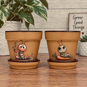 May include: Two tan terracotta plant pots with matching saucers and dark brown rims. The left pot features a painted pink caterpillar holding a strawberry, while the right pot displays a green caterpillar resting on a large orange carrot.