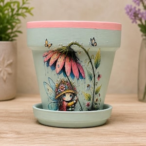 May include: A light mint green ceramic plant pot with a matching saucer, featuring a pink rim. The exterior displays a detailed illustration of a small fairy with wings under a large pink flower, accompanied by two small butterflies and green foliage.
