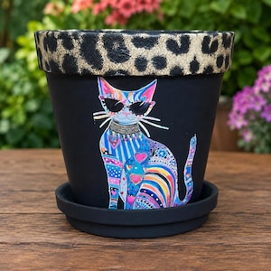 May include: A matte black terracotta plant pot with a matching saucer, featuring a gold and black leopard print rim and a colorful, stylized cat illustration wearing sunglasses and a collar on the front surface of the container.