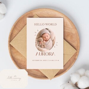 Op de afbeelding: Geboortekaart met de tekst "HELLO WORLD" en de naam "AURORA". De kaart toont een foto van een pasgeboren baby gewikkeld in een deken, met een teddybeer. De kaart ligt op een houten bord met een kraft papieren envelop.