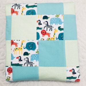 May include: A patchwork baby quilt with a blue and green color scheme. The quilt is made of squares of different fabrics, some with a white background and colorful animal prints, and some with solid colors. The quilt is perfect for a baby's nursery.