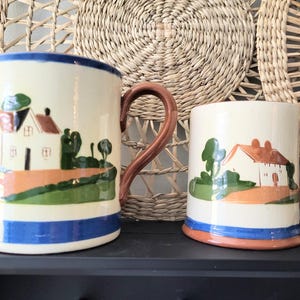 May include: Two ceramic mugs with a cream base, each decorated with a hand-painted rural scene of a house, trees, and a road. The mugs have brown handles and are trimmed with blue and orange bands. The mugs are displayed on a dark surface.