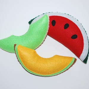 May include: Three felt fruit slices: a red watermelon wedge with black seeds, a green honeydew melon slice, and a yellow cantaloupe slice. The watermelon has a white rind and green outer edge. The other slices have brown edges.