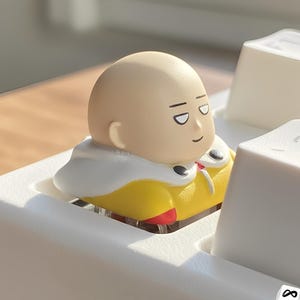 Könnte beinhalten: Eine individuelle Tastenkappe mit einem glatzköpfigen Charakter in einem gelben Anzug und weißem Umhang. Die Tastenkappe ist für eine mechanische Tastatur konzipiert. Der Charakter hat einen neutralen Gesichtsausdruck.