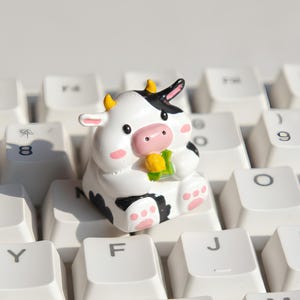 Könnte beinhalten: Ein schwarz-weißes Kuh-förmiges Keycap mit rosa Akzenten, das auf einer weißen Tastatur sitzt. Die Kuh hält ein gelbes Objekt. Die Tastaturtasten sind weiß mit schwarzer Beschriftung.