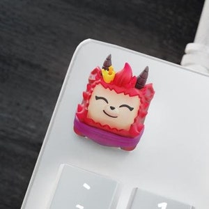 Op de afbeelding: Drie rode en witte toetsen met een cartoon-personage ontwerp. De toetsen zijn kubusvormig en hebben een glimlachend gezicht met hoorns en een kroon. De toetsen bevinden zich op een wit toetsenbord.