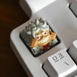 Op de afbeelding: Een wit toetsenbord met een klein, wit en oranje beeldje van een persoon met wit haar en een rode mond. Het beeldje staat op de toets met het label "/" en de toetsen eromheen zijn gelabeld "1!" en "FPS".