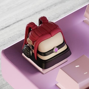 Può includere: Un keycap personalizzato raffigurante un personaggio femminile stilizzato con capelli rossi a codini. Il keycap ha un design nero e beige con una gemma viola, posto su una tastiera rosa.