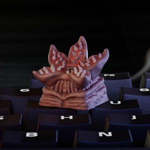 May include: A Demogorgon-themed keycap, featuring a detailed, multi-layered design with a reddish-pink flower-like head and sharp white teeth. The keycap is set on a dark keyboard, showcasing its unique, monster-inspired aesthetic.