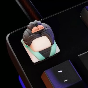 May include: A black and green keycap with a cartoon character wearing a green and white outfit. The character has black hair and a pink headband.