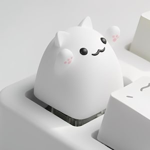 May include: A white cat-shaped keycap with black eyes and a smiling mouth sits on a white keyboard. The cat's paws are pink. The keyboard keys are white with black lettering. A fun and unique keyboard accessory.