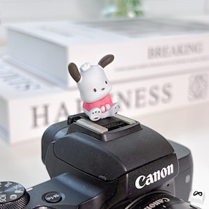 May include: A small, white and pink cartoon character figurine with brown ears sits atop a black Canon camera. The figurine is wearing a pink shirt. The camera has the Canon logo in white.