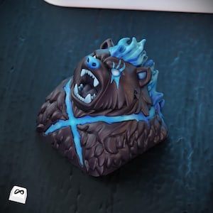 May include: A brown bear-shaped artisan keycap with blue accents. The bear has an open mouth, showing white teeth, and a blue cross design. The keycap has a textured, detailed design and a blue flame-like hair. The keycap is on a dark blue surface.