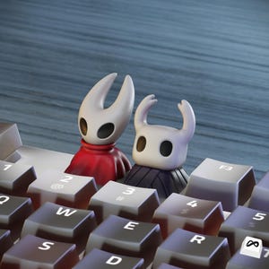 Puede incluir: Dos figuras estilizadas, una roja y otra negra, están sobre un teclado de ordenador. Las figuras tienen cabezas blancas con rasgos negros en forma de ojos. Las teclas del teclado son grises con letras blancas.