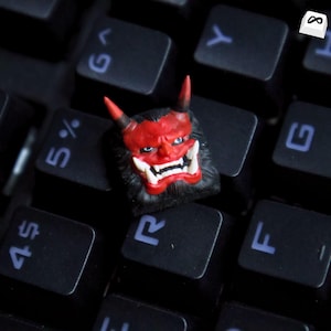Pode incluir: Uma tecla de teclado personalizada com um design de máscara Oni vermelho e preto. A tecla apresenta um rosto detalhado com dentes brancos, olhos azuis e chifres vermelhos. Em um teclado preto.