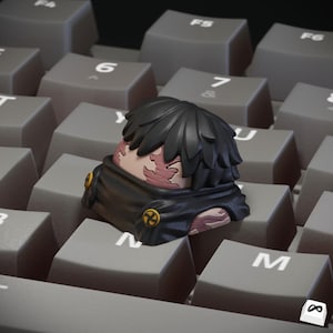 Può includere: Un copritasto personalizzato con un design di personaggio con capelli neri e una sciarpa nera. Il copritasto è posizionato su una tastiera grigia, con il viso del personaggio che mostra pelle rosa e segni di ustioni. Il copritasto ha un design unico.