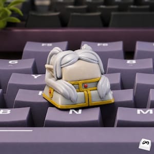 Op de afbeelding: Een op maat gemaakte artisan keycap met een gestileerd elf-ontwerp. De keycap heeft een lichtgrijze kop en armen, met een gele en gouden outfit. Het rust op een paars toetsenbord, met zichtbare letters en cijfers.