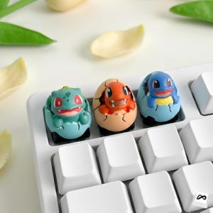May include: Three colorful, custom keycaps featuring Pokémon characters emerging from cracked eggs. The keycaps are green, orange, and blue, and are set on a white keyboard. The background includes white petals and green leaves.